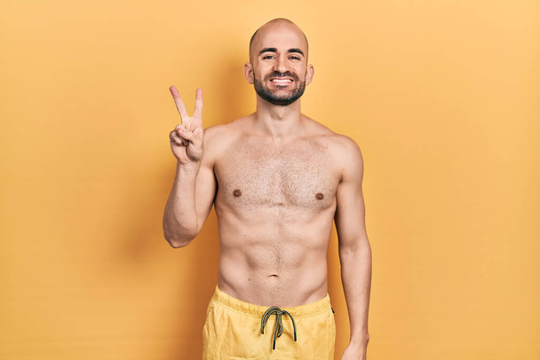 Young bald man wearing swimwear showing and pointing up with fingers number two while smiling confident and happy.  - Photo, Image