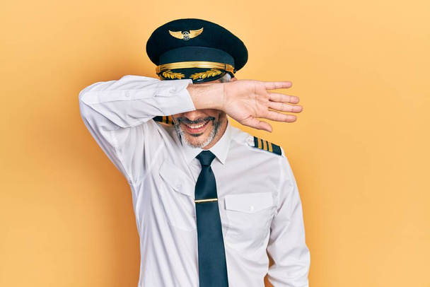 Charmant homme d'âge moyen aux cheveux gris portant l'uniforme de pilote d'avion couvrant les yeux avec le bras souriant gai et drôle. concept aveugle.  - Photo, image