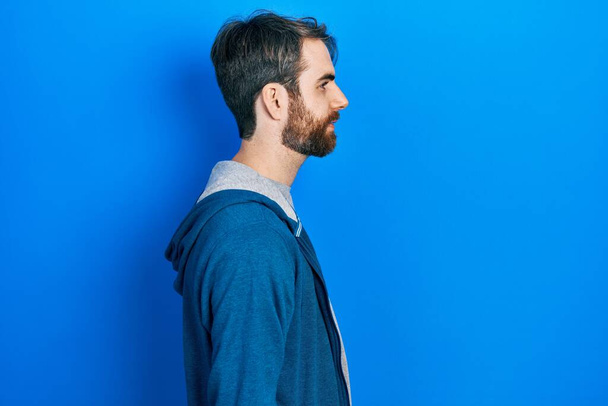 Homme caucasien avec barbe portant sweat-shirt décontracté regardant vers le côté, pose de profil relax avec visage naturel avec sourire confiant.  - Photo, image