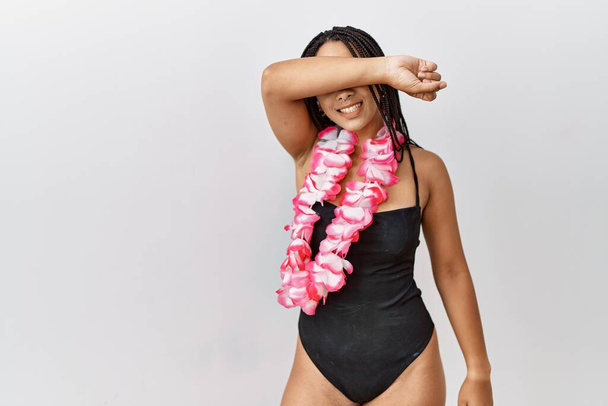 Junge afrikanisch-amerikanische Frau in Badeanzug und hawaiianischem Blei bedeckt Augen mit Arm lächelt fröhlich und lustig. Blindes Konzept.  - Foto, Bild