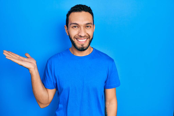 Латиноамериканец с бородой в повседневной синей футболке улыбается веселым подарком и указывает пальцем на камеру.  - Фото, изображение