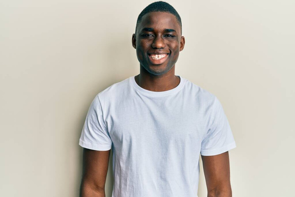 Молодой африканский американец в белой футболке выглядит позитивно и счастливо стоя и улыбаясь с уверенной улыбкой, показывая зубы  - Фото, изображение