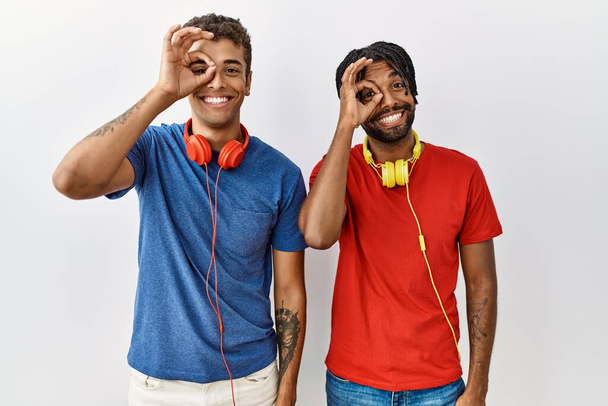 Молодые испаноязычные братья стоят на изолированном фоне в наушниках и делают все хорошо жесты с улыбкой на руке, глаза смотрят сквозь пальцы с счастливым лицом.  - Фото, изображение
