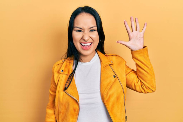 Красивая латиноамериканка с пирсингом носа в желтой кожаной куртке показывает и указывает пальцами номер пять, улыбаясь уверенно и счастливо.  - Фото, изображение