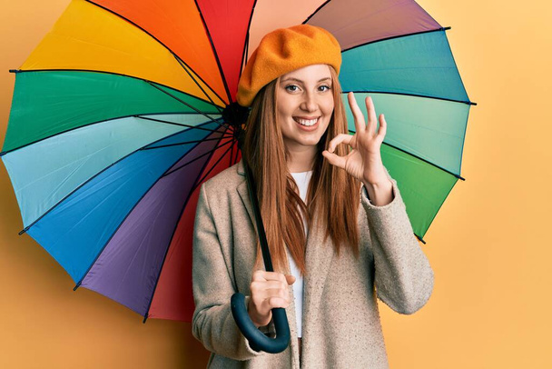Fransız tarzı giyinen genç İrlandalı kadın elinde renkli bir şemsiye tutuyor parmaklarıyla işaret veriyor, gülümsüyor ve mükemmel bir sembol oluşturuyor.  - Fotoğraf, Görsel