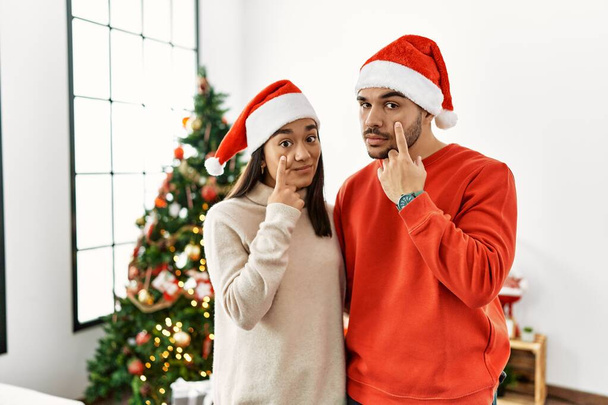 Junges hispanisches Paar, das am Weihnachtsbaum steht und auf das Auge zeigt, das dich beobachtet, Geste, misstrauischer Ausdruck  - Foto, Bild