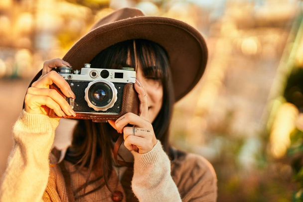Mujer morena con sombrero de invierno sonriendo usando una cámara vintage en el parque - Foto, imagen