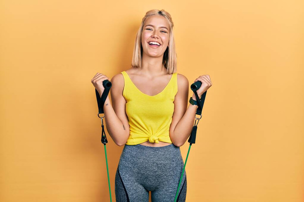 Mooie blonde vrouw training arm weerstand met elastische armbanden glimlachen en hard lachen hardop omdat grappige gekke grap.  - Foto, afbeelding