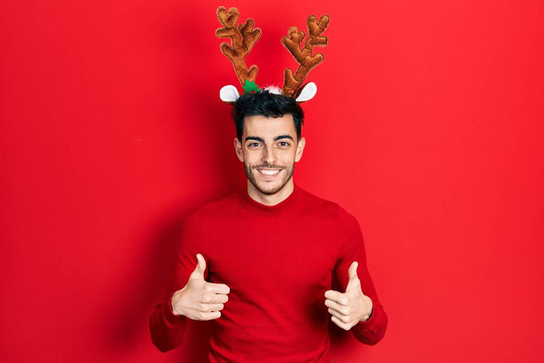 Young hispanic man wearing cute christmas reindeer horns success sign doing positive gesture with hand, thumbs up smiling and happy. cheerful expression and winner gesture.  - Photo, Image
