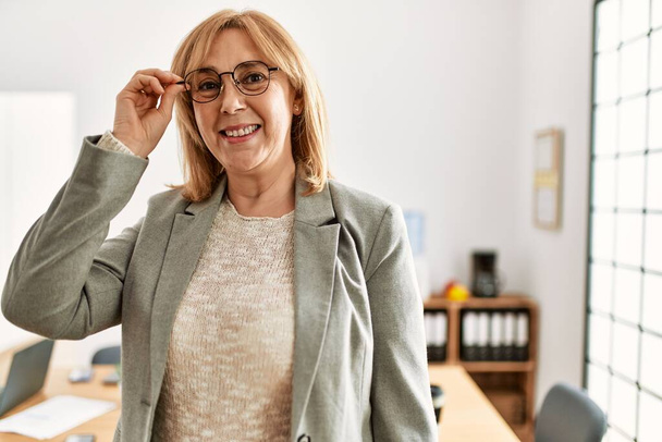 Mulher de negócios de meia idade sorrindo feliz segurando óculos no escritório. - Foto, Imagem