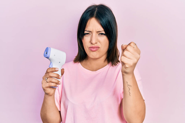 Mujer hispana joven sosteniendo termómetro molesto y frustrado gritando con ira, gritando loco de ira y la mano levantada  - Foto, Imagen