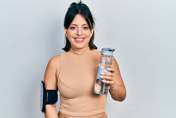 Молодая латиноамериканка в спортивной одежде пьет бутылку воды, выглядит позитивно и счастливо стоя и улыбаясь с уверенной улыбкой, показывая зубы  - Фото, изображение
