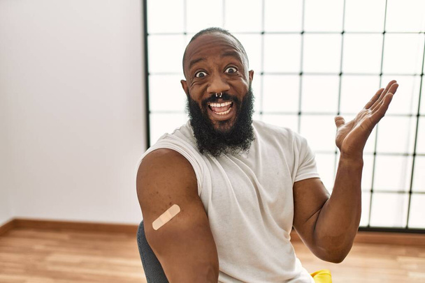 African american man getting vaccine showing arm with band aid celebrating victory with happy smile and winner expression with raised hands  - Foto, Imagen