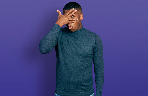 Молодой черный мужчина в обычном свитере подглядывает рукой в лицо и глаза, испуганно глядя сквозь пальцы.  - Фото, изображение