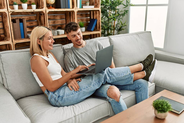 Νεαρό καυκάσιο ζευγάρι χαμογελά χαρούμενος χρησιμοποιώντας φορητό υπολογιστή κάθεται στον καναπέ στο σπίτι. - Φωτογραφία, εικόνα