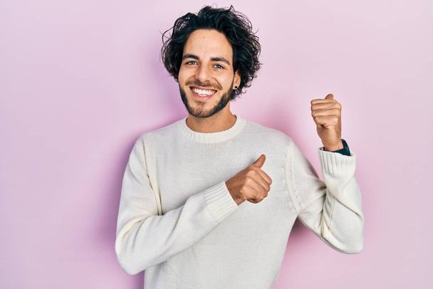 Schöner hispanischer Mann in lässigem weißen Pullover, der mit erhobenem Daumen nach hinten zeigt und selbstbewusst lächelt  - Foto, Bild