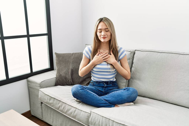 Азиатская молодая женщина, сидящая дома на диване, улыбаясь руками на груди с закрытыми глазами и благодарным жестом на лице. концепция здоровья.  - Фото, изображение