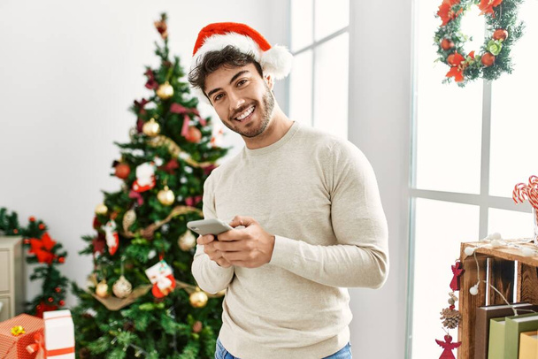 Giovane uomo ispanico indossa cappello di Natale utilizzando smartphone a casa. - Foto, immagini