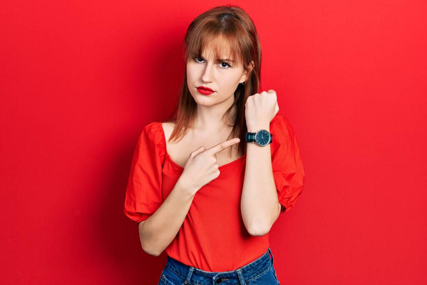 Rudowłosa młoda kobieta w czerwonej koszuli w pośpiechu wskazując czas, niecierpliwość, patrząc w kamerę z miłą ekspresją  - Zdjęcie, obraz