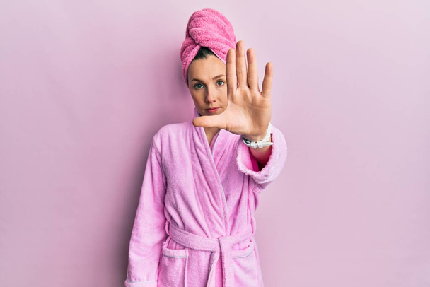 Young blonde woman wearing shower towel cap and bathrobe doing stop sing with palm of the hand. warning expression with negative and serious gesture on the face.  - Photo, Image