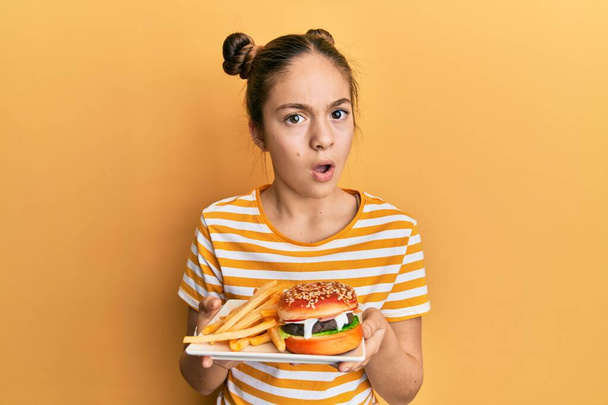Beautiful brunette little girl eating a tasty classic burger with fries in shock face, looking skeptical and sarcastic, surprised with open mouth  - Photo, Image