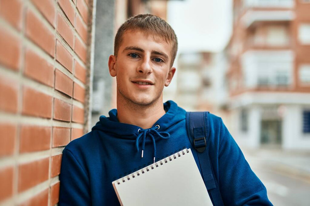 Young blond student smiling happy holding notebook at the university. - 写真・画像