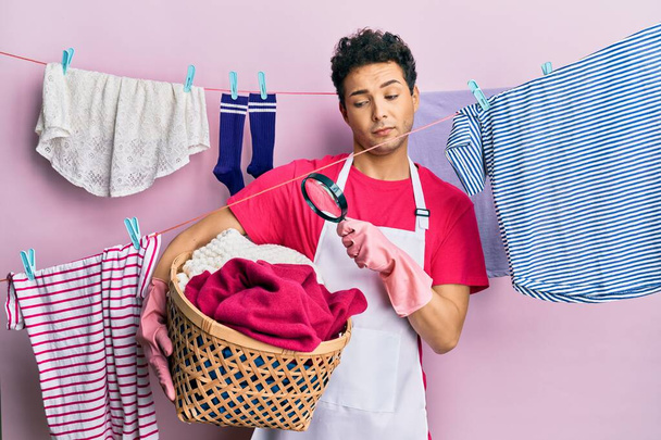 Bonito hispânico fazendo lavandaria segurando cesta de vime à procura de uma mancha no rosto de choque, olhando cético e sarcástico, surpreso com a boca aberta  - Foto, Imagem