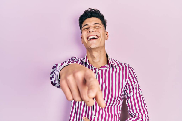 Mladý hispánec v neformálním oblečení, směje se ti, ukazuje prstem na kameru s rukou přes tělo, výraz studu  - Fotografie, Obrázek