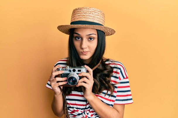 Mulher hispânica jovem usando chapéu de verão segurando câmera vintage cético e nervoso, franzindo a testa chateado por causa do problema. pessoa negativa.  - Foto, Imagem