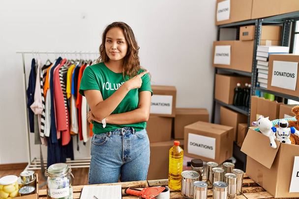 Beautiful hispanic woman wearing volunteer t shirt at donations stand pointing with hand finger to the side showing advertisement, serious and calm face  - Foto, Imagen