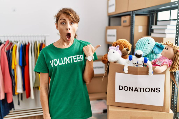 Mooie blanke vrouw draagt vrijwilliger t-shirt bij donaties staan verrast wijzend met de hand vinger naar de zijkant, open mond verbaasd uitdrukking.  - Foto, afbeelding