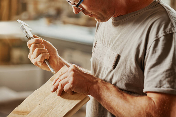 Menuisier prenant une mesure d'une planche de bois avec un étrier dans un gros plan sur ses mains et l'outil dans un grand atelier - Photo, image