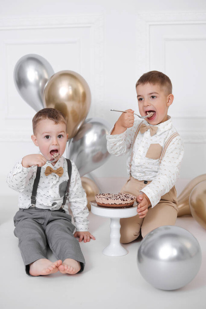 Dos chicos celebrando su cumpleaños, los niños tienen una fiesta del día B. Tarta de cumpleaños con velas y globos. Niños felices comiendo pastel, celebración, interior minimalista blanco. - Foto, imagen