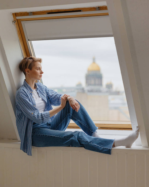 ikkunalaudalla istuu nuori ja kaunis eurooppalainen nainen, jolla on lyhyet vaaleat hiukset.  - Valokuva, kuva