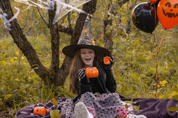 Funny child girl in a witch costume for Halloween holds a mug in the form of a pumpkin. - Fotó, kép