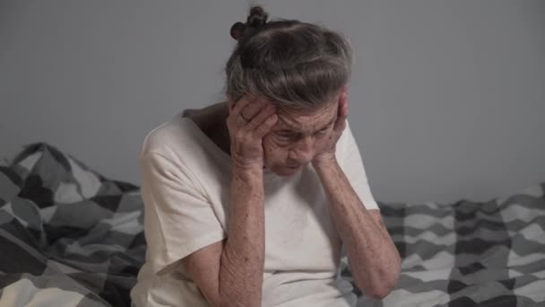 Старшая белая женщина с седыми волосами 90 лет держит голову, чувствует головную боль и высокое кровяное давление, проверяет пульс, пульс, сидя на кровати. Гипертония у пожилой женщины в доме престарелых - Кадры, видео