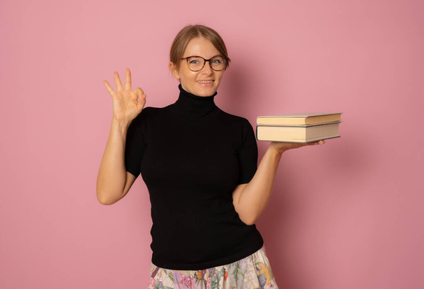 Photo de jeune belle fille souriante positive gaie dans des lunettes avec pile de livres isolés sur fond de couleur rose - Photo, image