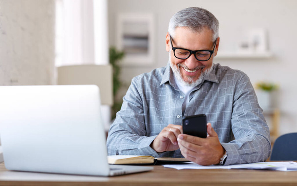 Komea hymyilevä vanhempi mies, jolla on lasit matkapuhelimella, kun hän istuu kodikas työpaikallaan kannettavan tietokoneen kanssa kotona, eläkkeellä oleva mies keskustelee ystävien kanssa sosiaalisessa mediassa, kirjoittamalla älypuhelimeen - Valokuva, kuva