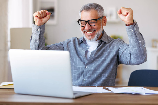Excited senior man celebrating success, getting good exam results during distant education, raising hands with clenched fists and looking at laptop with happy face expression while sitting at desk - Фото, изображение
