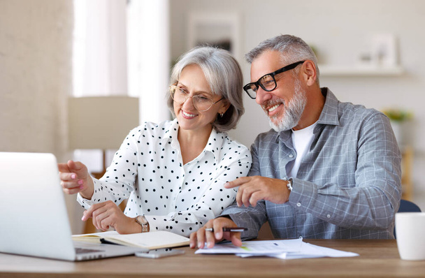 Старшая семейная пара учится онлайн вместе, мужчина и женщина на пенсии смотрят на ноутбук и обсуждают что-то, сидя за столом дома, наслаждаясь дистанционным образованием на пенсии - Фото, изображение