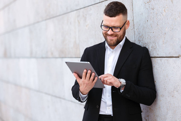 Positieve vrolijke zakenman in stijlvol pak met digitale tablet terwijl hij buiten tegen een betonnen muur staat, tevreden kantoormedewerker die e-mail typt op modern elektronisch apparaat - Foto, afbeelding