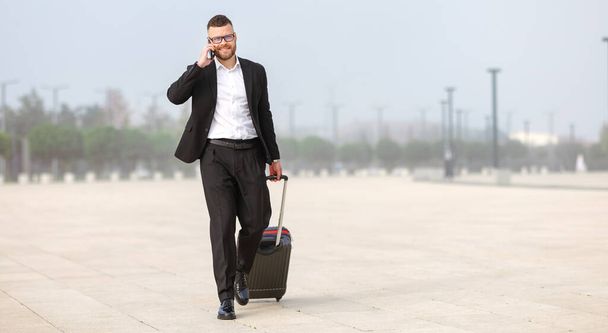Teljes hossza fiatal magabiztos üzletember formális fekete öltöny húzza bőrönd poggyász és telefonon leszállás után a repülőtéren, hívja a taxit. Üzleti út koncepciója - Fotó, kép