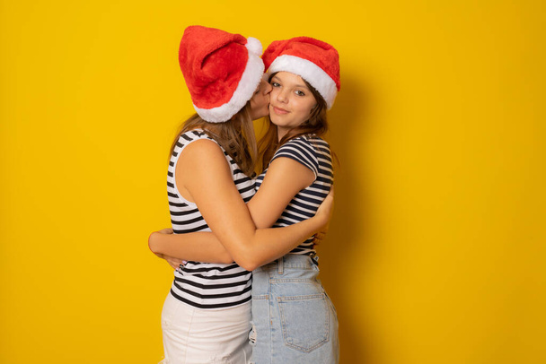 Matka líbá svou dceru v Santa klobouku přes žluté pozadí. Vánoční koncept. - Fotografie, Obrázek