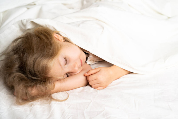 Una rubiecita está durmiendo dulcemente en una cama con ropa interior blanca. espacio para el texto. El sueño sano del niño. - Foto, imagen