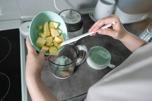Gros plan sur la main d'une femme inconnue tenant un bol avec des tranches de pomme de terre coupées verser au mélangeur pour faire un repas dans la cuisine - Photo, image