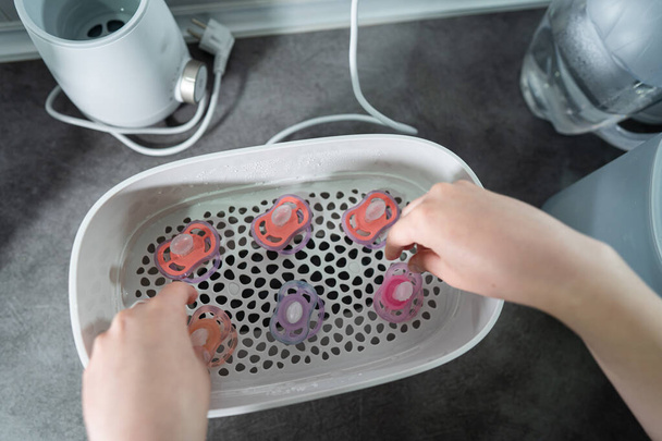 Nahaufnahme einer unbekannten Frau, die Hand voller Schnuller über einem elektrischen Dampfdesinfektionsbehälter auf der Küchentheke hält - Foto, Bild