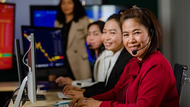 Группа азиатских женщин-операторов обслуживания клиентов носит микрофонные наушники сидеть улыбаясь перед ежедневным графиком монитор компьютера монитор говорить и торговать купить акции рынка онлайн с клиентом. - Фото, изображение