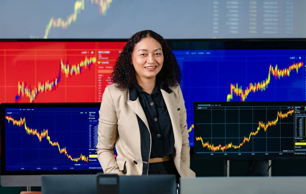 女性の成功したプロのブローカートレーダー投資家は、株式とビットコイン暗号通貨グラフ分析とフロントコンピュータモニター画面のカメラでパンツポケットルックで手を入れて笑顔スタンド. - 写真・画像