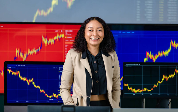 女性の成功したプロのブローカートレーダー投資家は、株式とビットコイン暗号通貨グラフ分析とフロントコンピュータモニター画面のカメラでパンツポケットルックで手を入れて笑顔スタンド. - 写真・画像