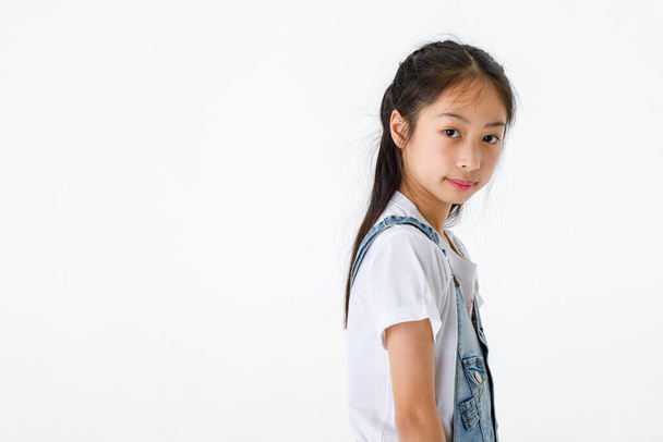 Portrait close up isolated studio shot of young pretty cute long black hair elementary girl in jeans denim skirt overalls standing smiling look at camera in front of white background with copy space. - Photo, Image